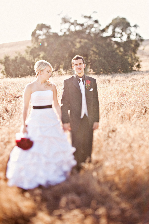 Bride and groom standing in an open field - Bride is wearing white ruffled ball gown style dress  with black sash carrying a red bouquet and groom is wearing black suit with white tie with red boutonniere -  photo by Orange County based wedding photographers Mark Brooke